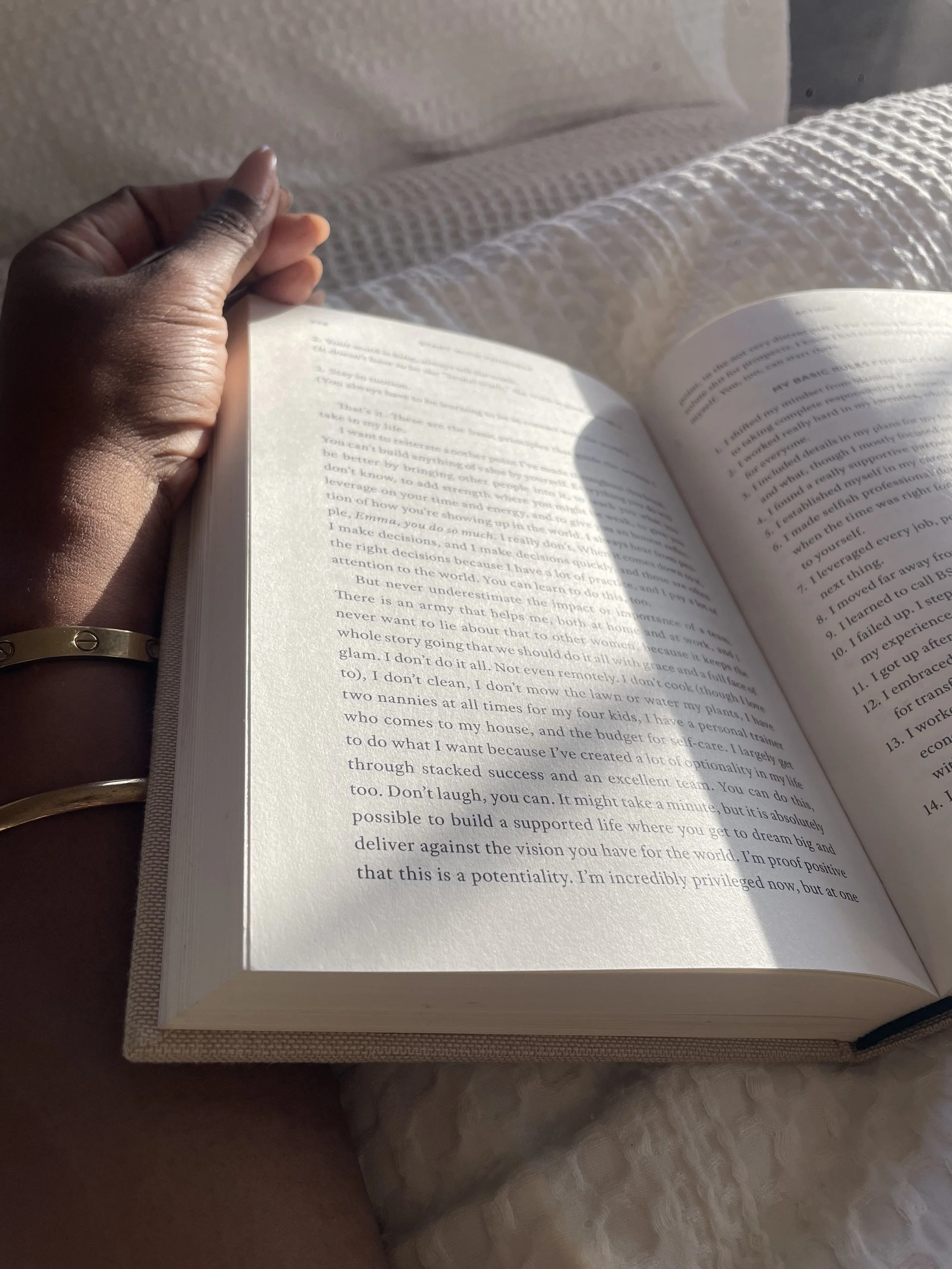 Hands holding open Emma Grede Start With Yourself book in morning light on white bedding
