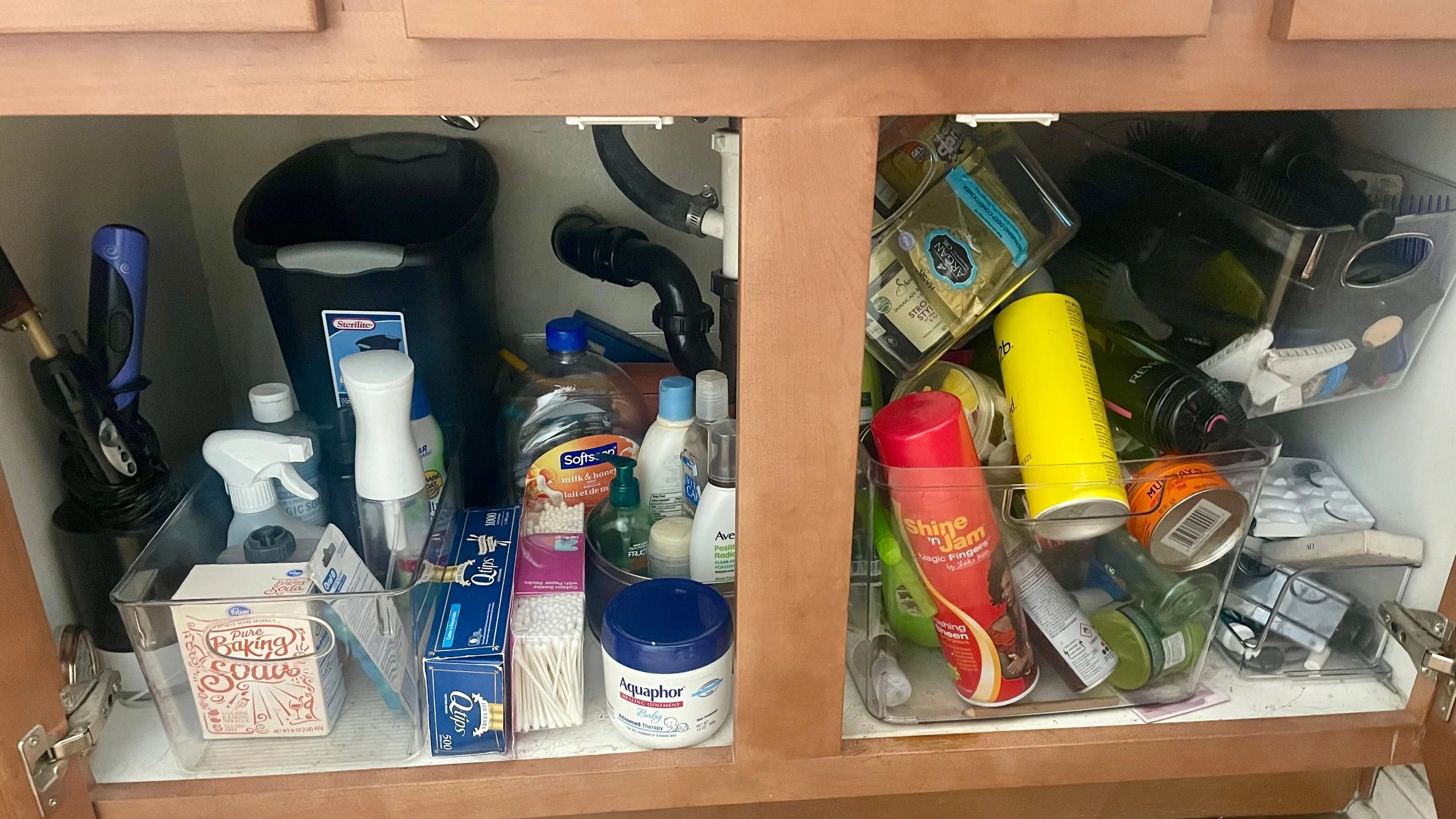 Cluttered under-sink bathroom cabinet with mismatched bins, loose products, hair tools, and no clear organization system.