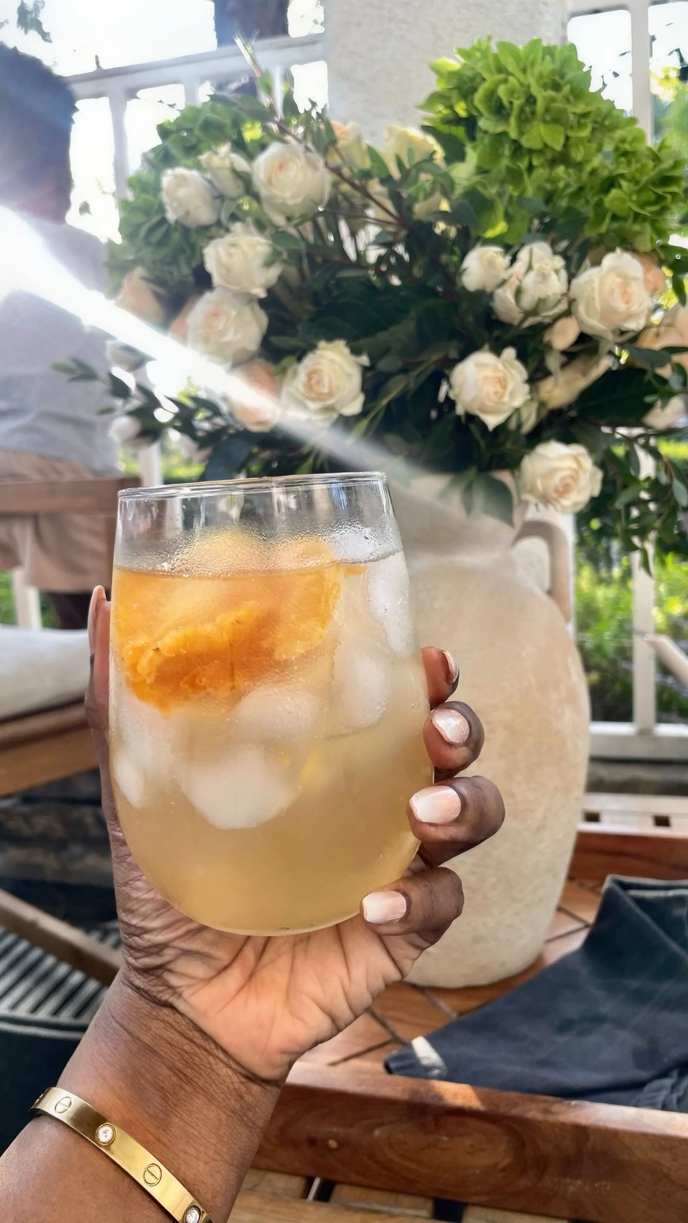 Hand holding a chilled iced green tea with pineapple and lemon on a sunlit patio with soft florals in the background