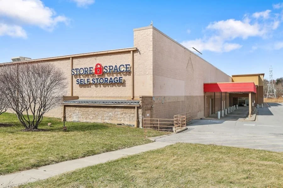 Empty parking lot and storefront of Store Space self-storage facility, with a beige and brick exterior, and a red awning over the entrance.