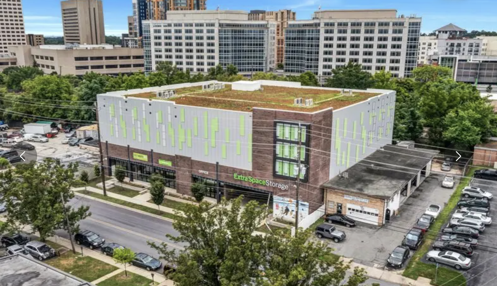 A multi-story building in an urban area with a green and white exterior and a green sign that reads 'Extra Space Storage'. The building has a parking lot with cars, trees lining the sidewalk, and high-rise buildings in the background.