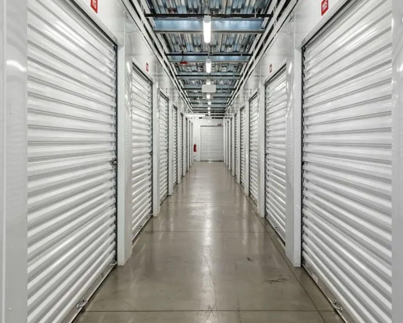 A corridor with multiple storage unit doors, all white, in a self-storage facility. The floor is concrete, and the ceiling has fluorescent lighting and metal pipes.