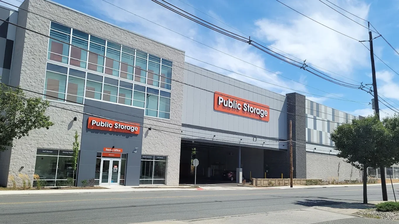 A large public storage facility with signs indicating 'Public Storage' on the building, a parking entrance, and windows on the upper floors, under a partly cloudy sky.