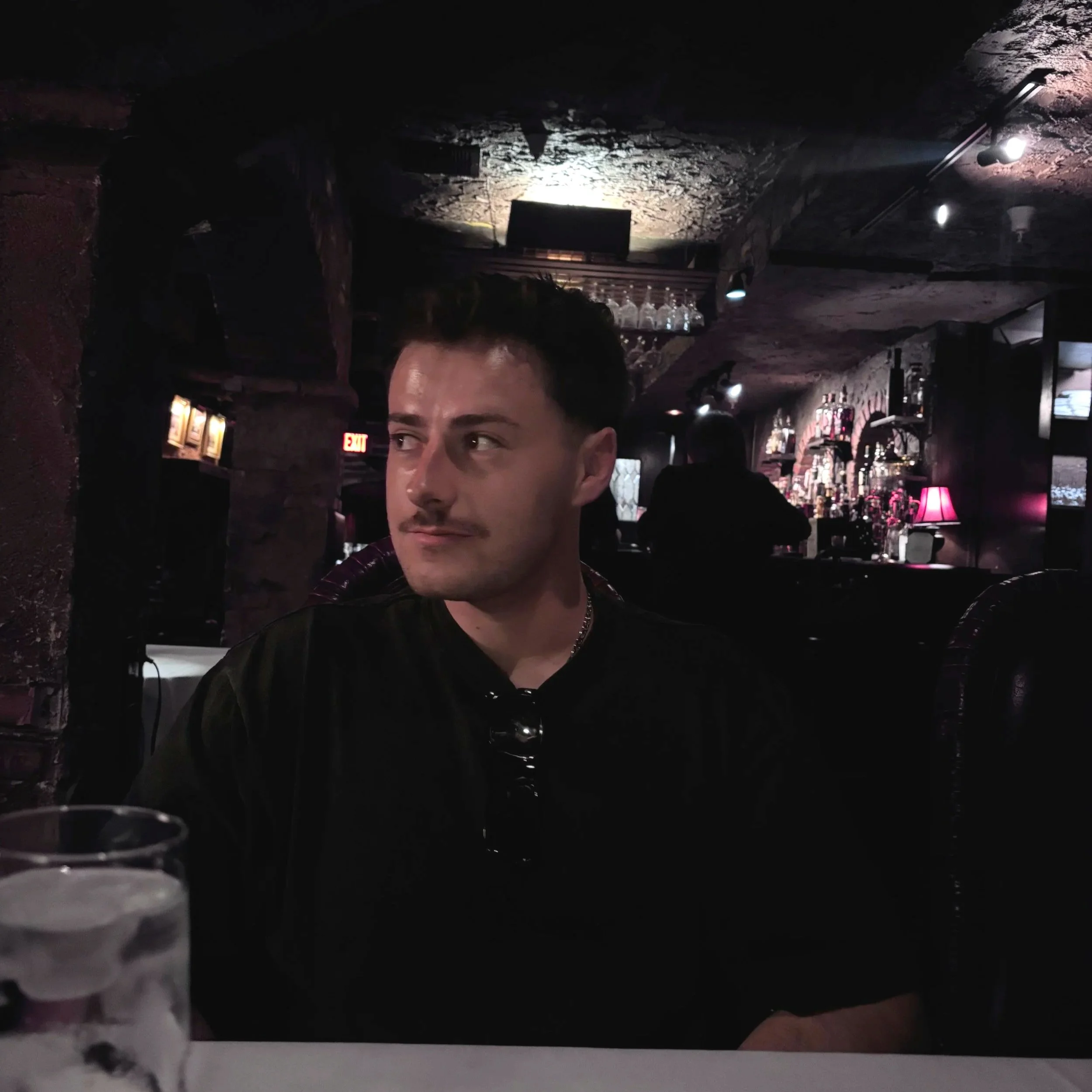 A person with dark hair and a light complexion in a dimly lit bar, with liquor bottles on shelves in the background and a bar counter in front.