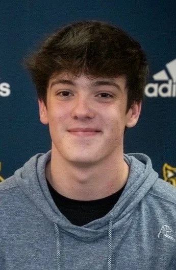 A young man with brown hair wearing a gray hoodie, smiling in front of a dark blue background with sports logos.