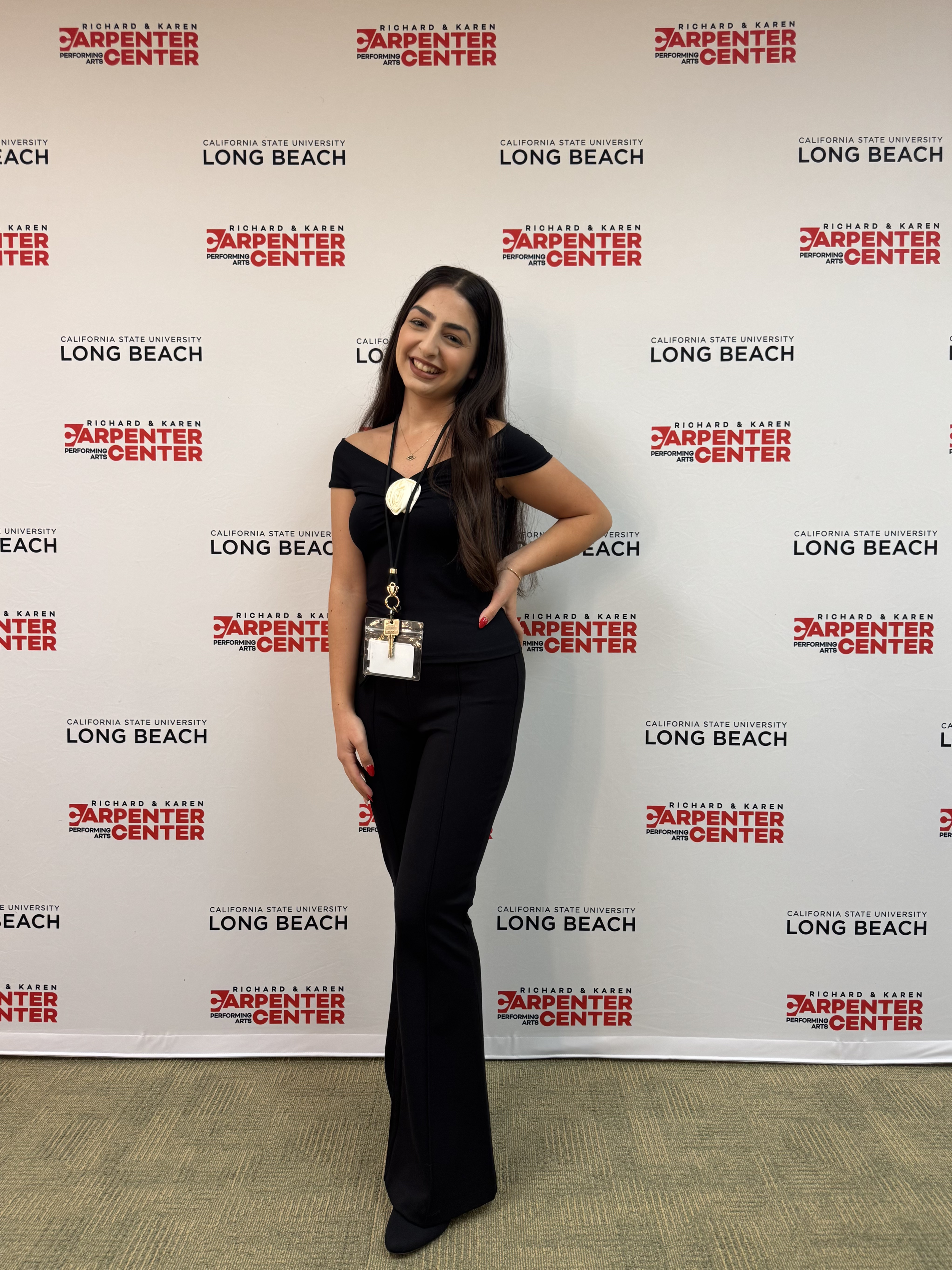 A woman with long dark hair wearing a black off-shoulder top and black pants, smiling and standing with her hand on her hip in front of a backdrop that reads 'Richard & Karen Carpenter Center Performing Arts California State University Long Beach'.