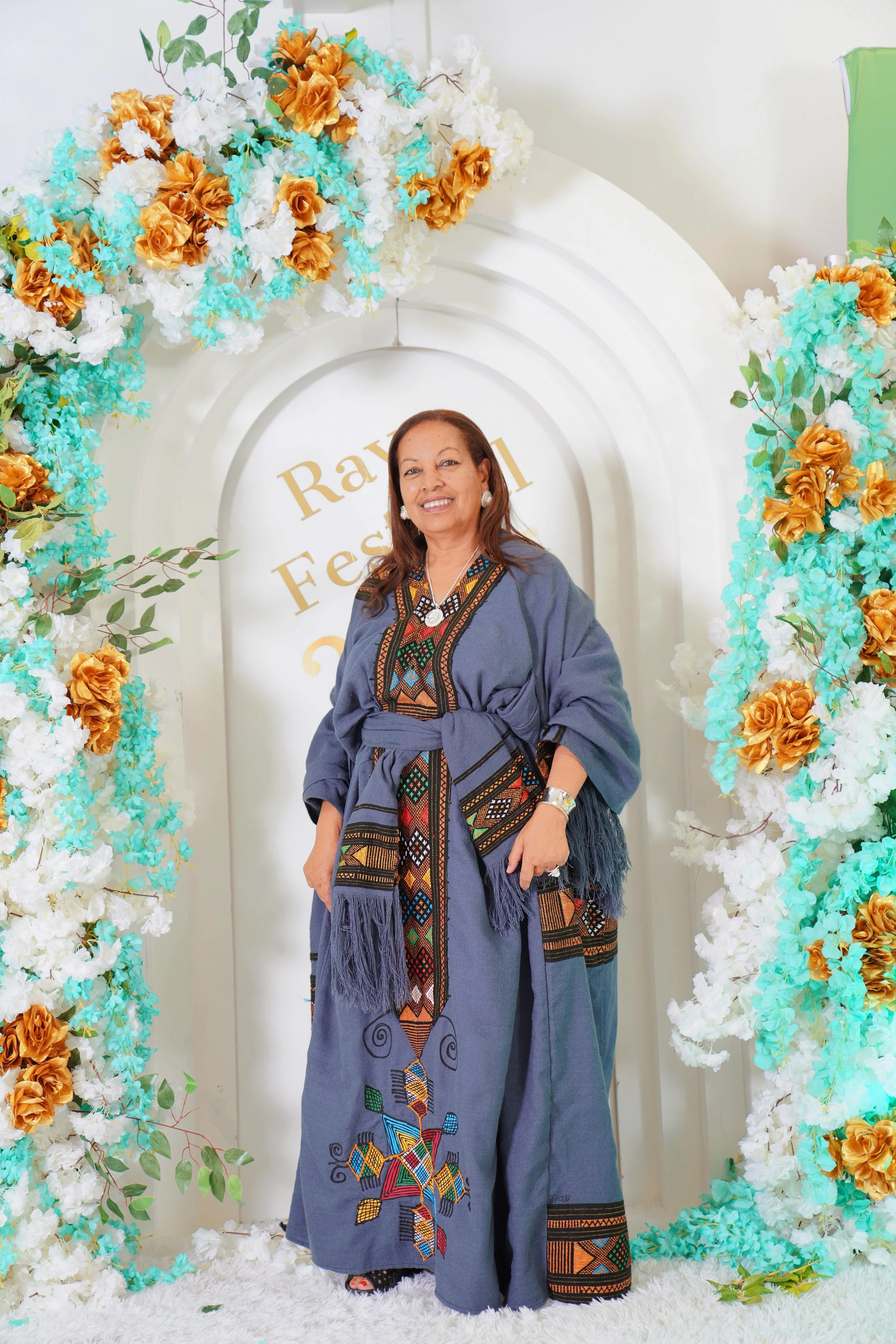 A smiling woman standing in front of a floral arch with white, orange, and turquoise flowers. She is wearing a long, patterned, blue dress with fringe details, and accessorized with earrings, bracelets, and a necklace. The background has a white wall