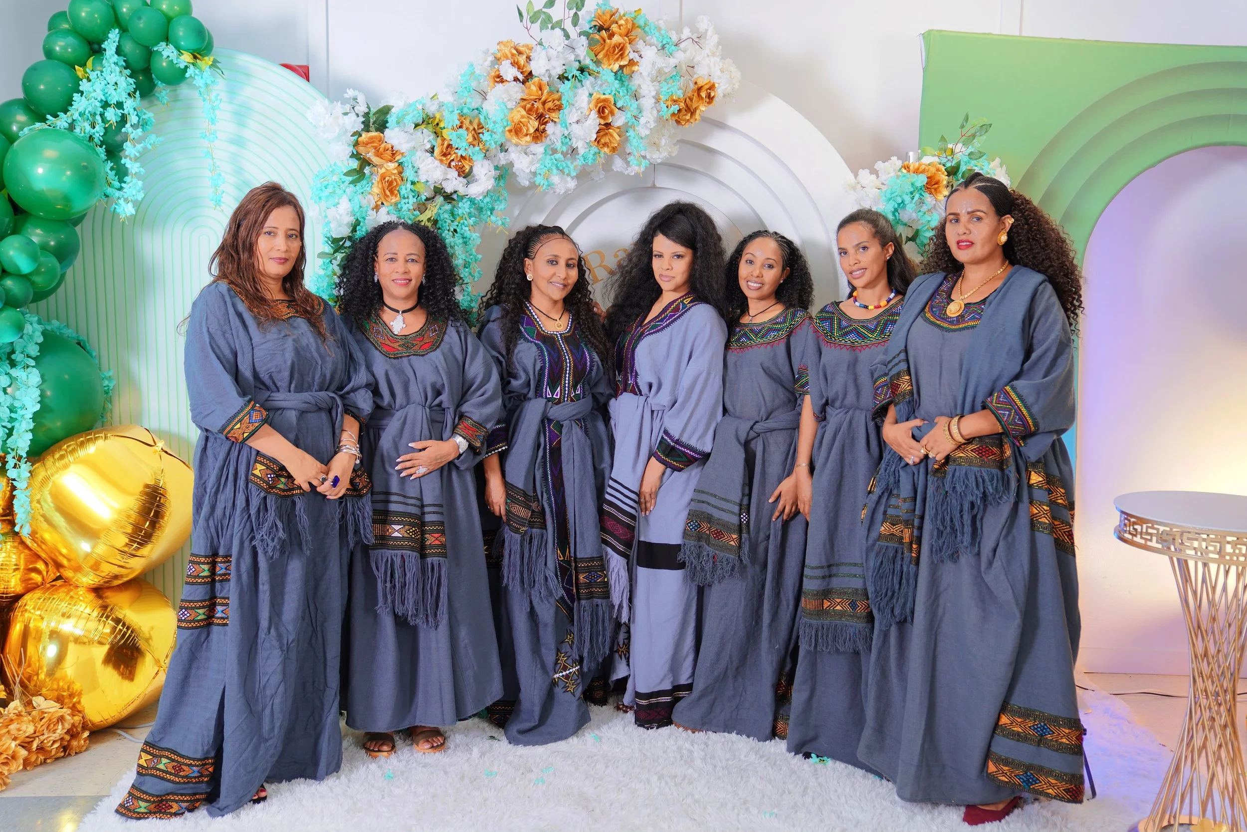 Six women dressed in matching traditional gray outfits with colorful embroidery, standing in front of a decorative backdrop with green and gold balloons and floral arrangements at a celebration or event.