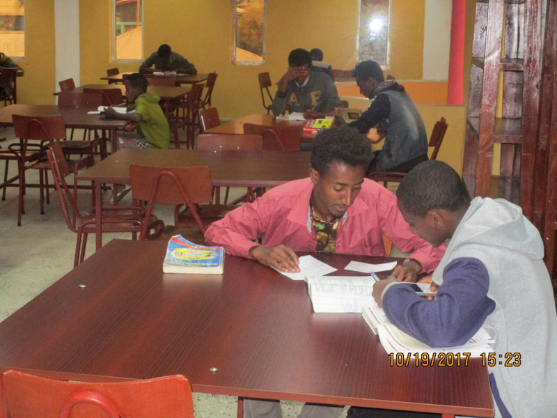 Students studying at tables in a library or study room, with books and notes on the tables.