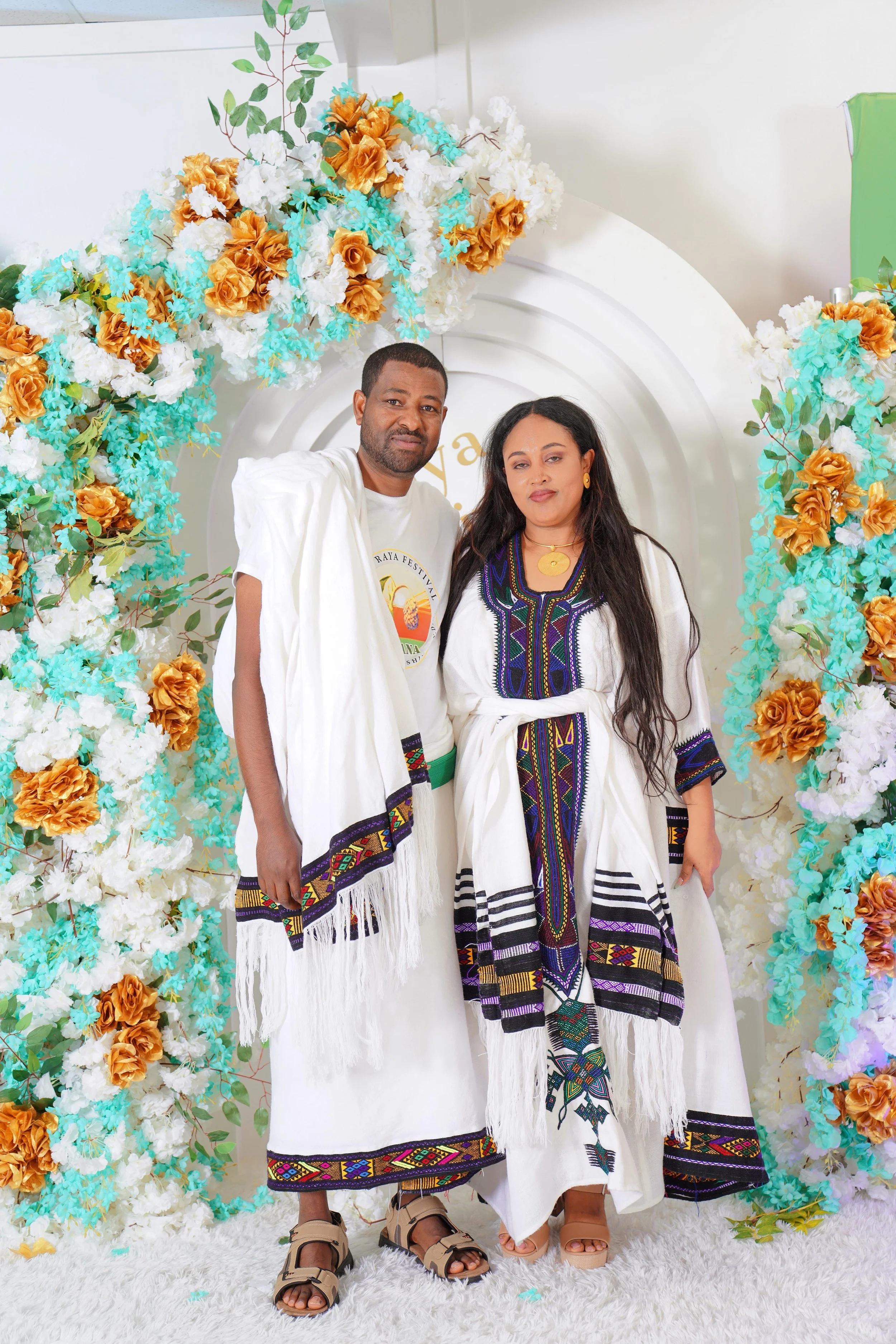 Two people wearing traditional embroidered clothing, standing in front of a floral arch with white, turquoise, and gold flowers, at a festive event.