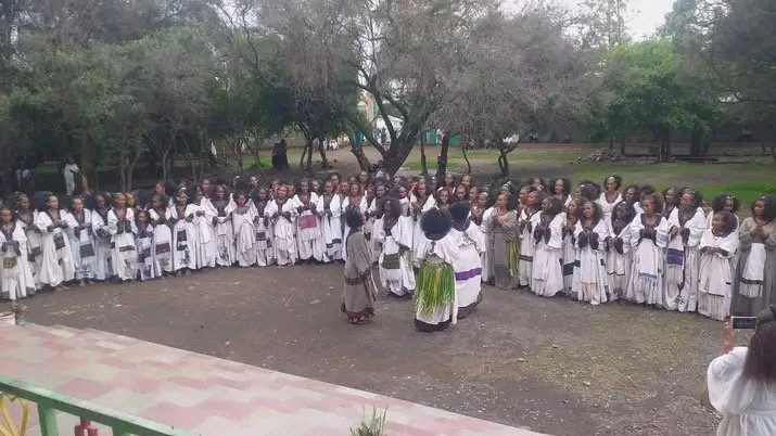 Group of children dressed in traditional clothing standing outdoors in a semi-circle, with some adults or elders in traditional robes in the center. Trees are in the background.