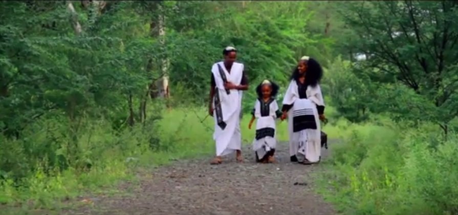 A family of three walking on a dirt trail through a lush green forest, dressed in traditional white and black clothing.
