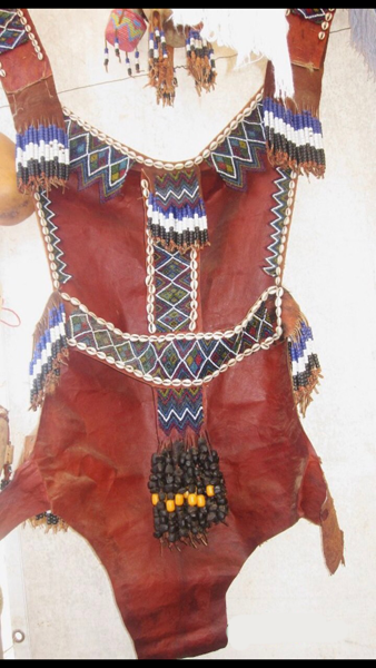 Traditional leather garment decorated with colorful beadwork and chains, featuring fringe and hanging ornaments.