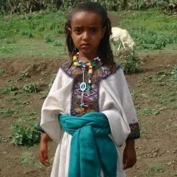 Young girl wearing traditional clothing with colorful beads and a turquoise sash, standing outdoors in a rural setting.