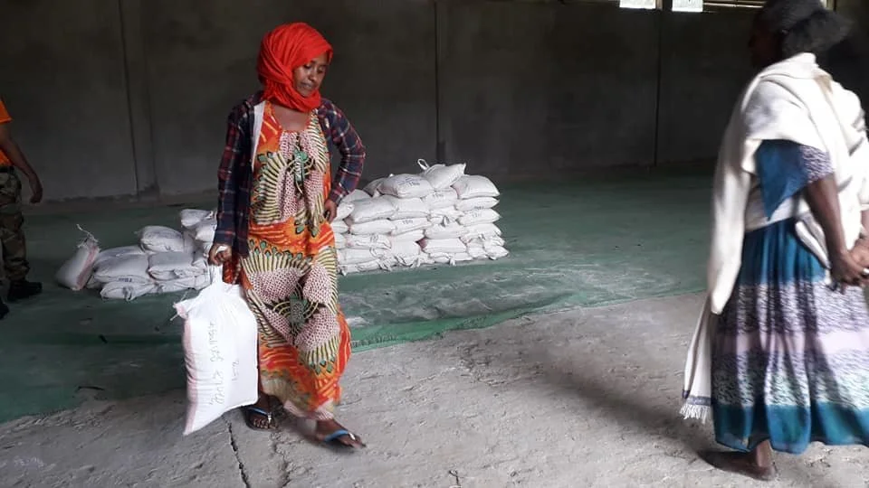 Two women in a warehouse, one carrying a sack of aid supplies, with more stacked behind them.