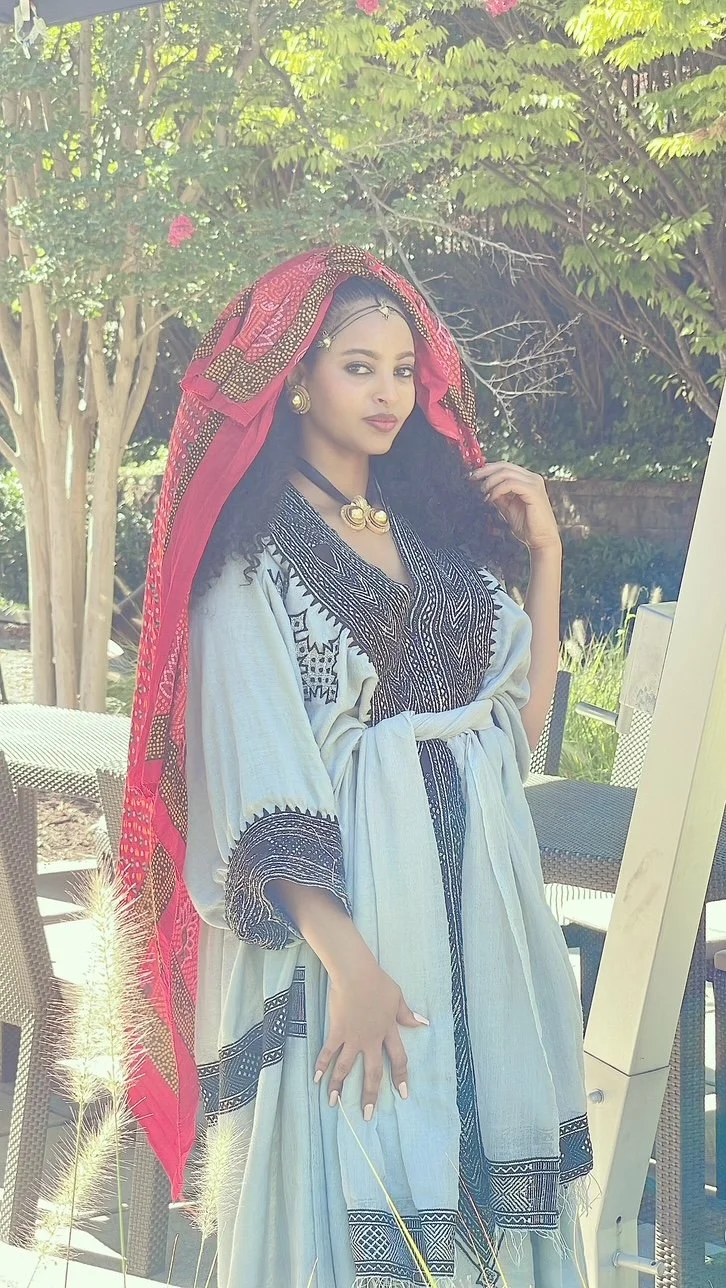 A woman dressed in traditional African attire, wearing a white and black embroidered dress with a red and gold head wrap, accessorized with gold jewelry, standing outdoors under green trees.