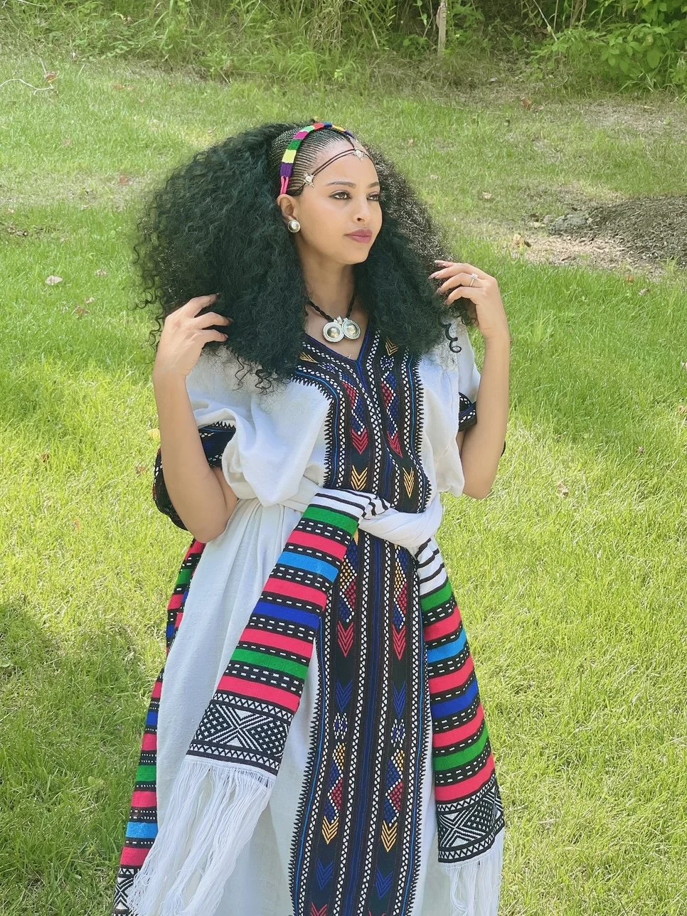 Woman standing outdoors on grass, wearing traditional African dress with colorful embroidery, black curly hair, headband, earrings, and necklace.