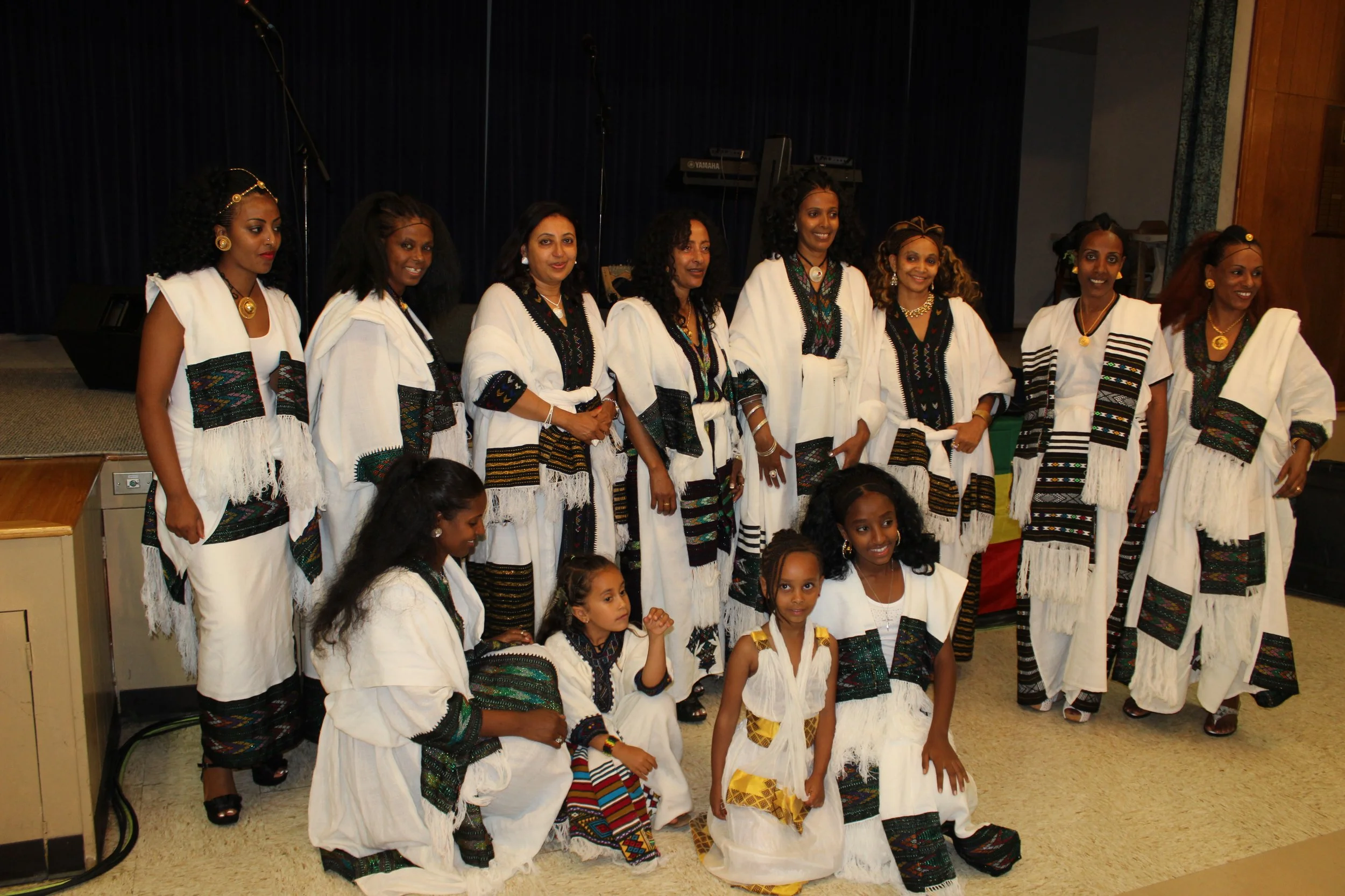 Group of women and children in traditional African clothing standing on a stage or platform inside a hall or auditorium.