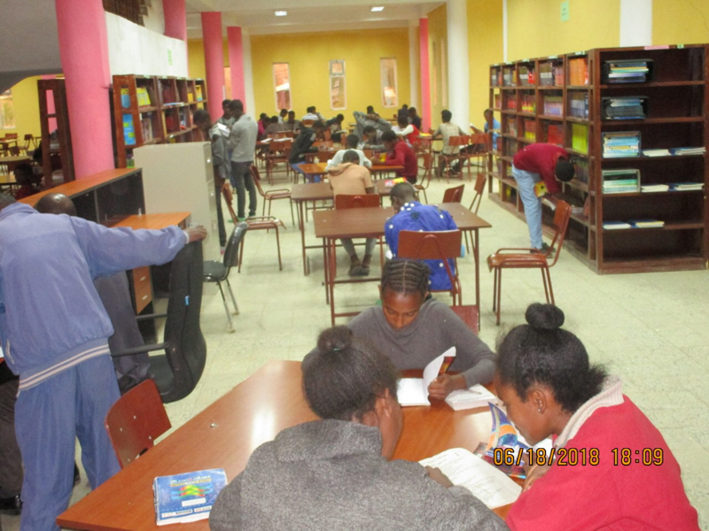 Students studying in a library, reading books at tables, with bookshelves in the background.