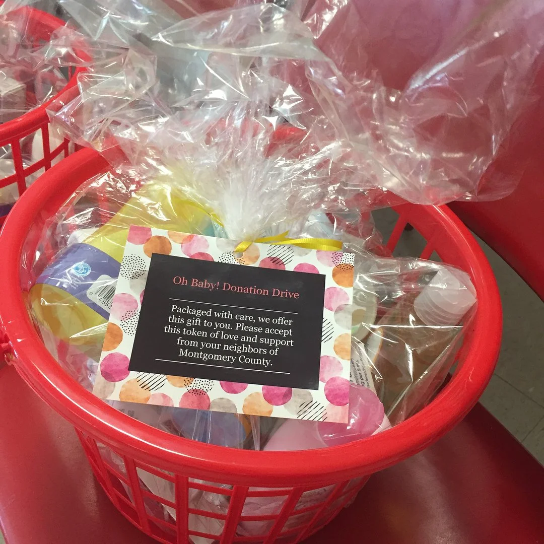 A red plastic basket filled with small gift packets wrapped in clear plastic, with a black and colorful note attached that reads, 'Oh Baby! Donation Drive. Packaged with care, we offer this gift to you. Please accept this token of love and support from your neighbors of Montgomery County.'