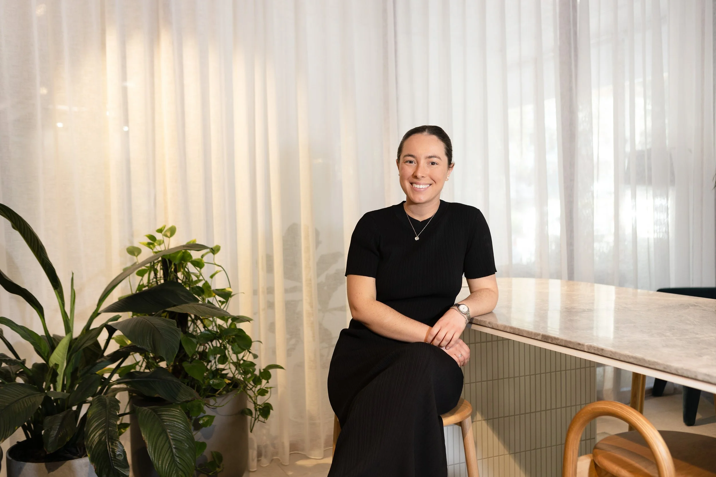 A woman sitting on a stool at a marble counter, smiling, wearing a black dress, and surrounded by green potted plants in a bright room with sheer curtains.