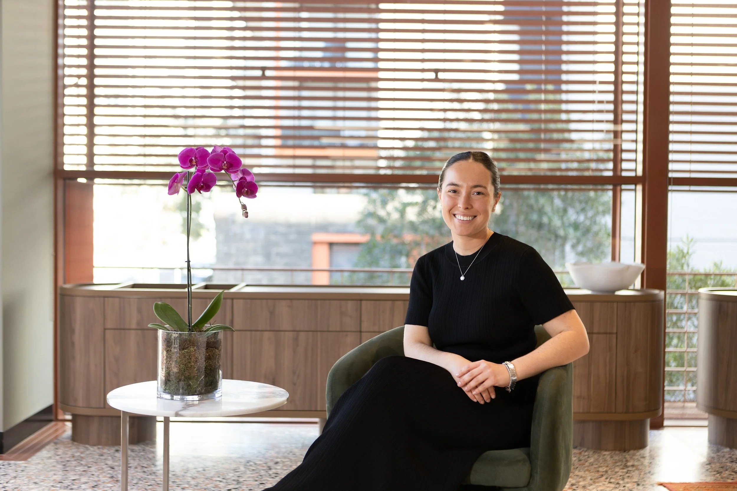 A woman in a black dress sits smiling on a green armchair in a room with large window blinds. A potted purple orchid is on a small white table next to her, and a wooden cabinet is in the background.
