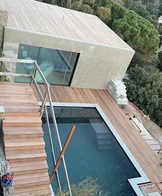 Modern house with a rooftop pool, wooden deck, and glass barriers, overlooking a green landscape.