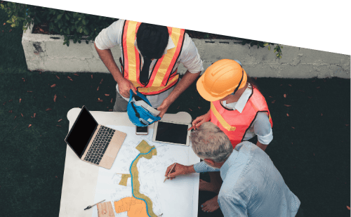 Two construction workers and a man in a blue shirt examining a large map on a table outdoors, with laptops and tablets nearby.