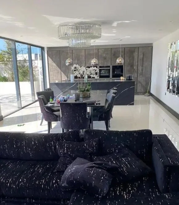 Modern kitchen and dining area with black and gray color scheme, featuring a black marble dining table, black chairs, a black sofa with pillows, and sleek gray cabinetry, illuminated by pendant lights and a large chandelier.
