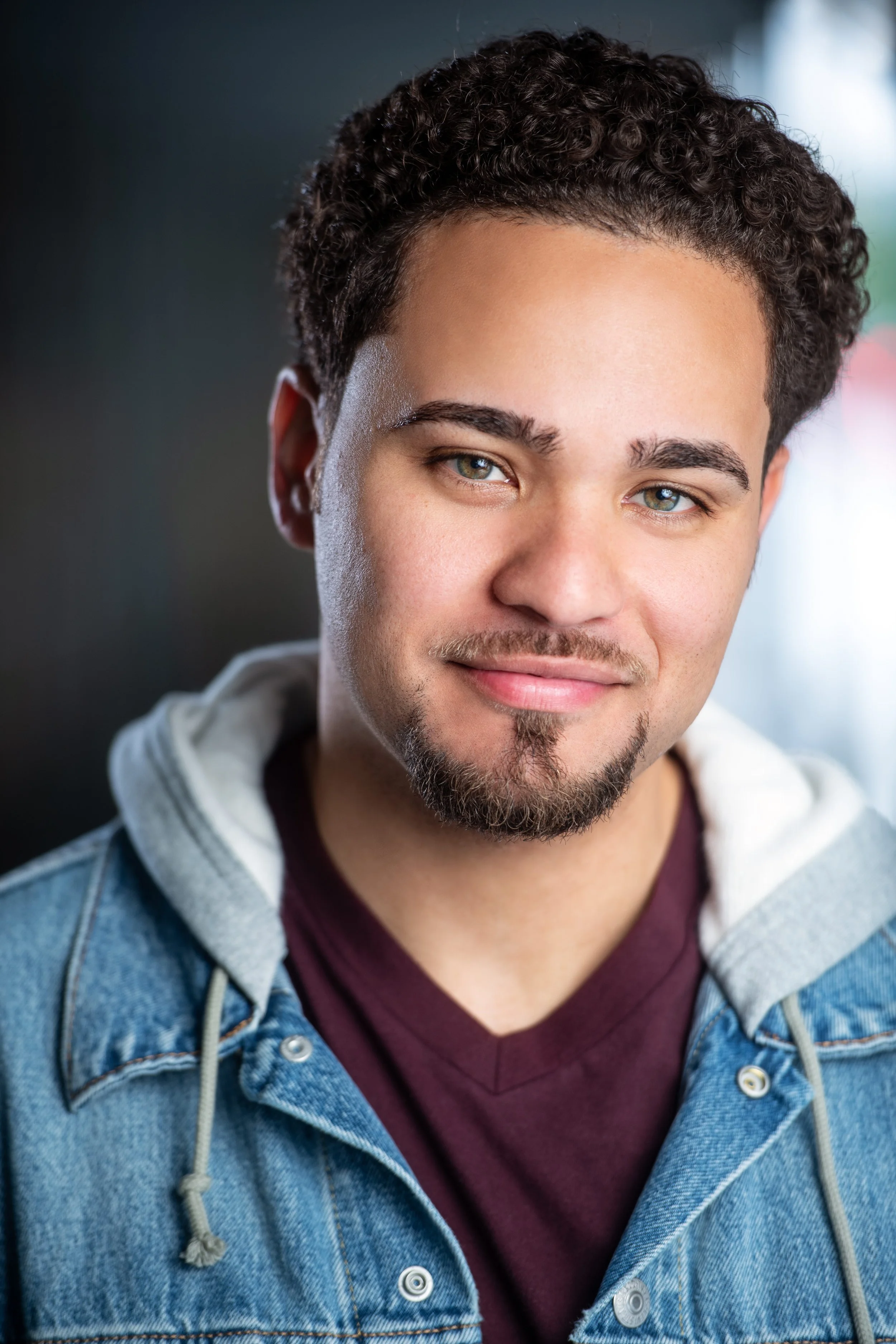 Close-up portrait of a young man with curly dark hair, hazel eyes, and a light beard, wearing a denim jacket over a maroon t-shirt, smiling slightly.