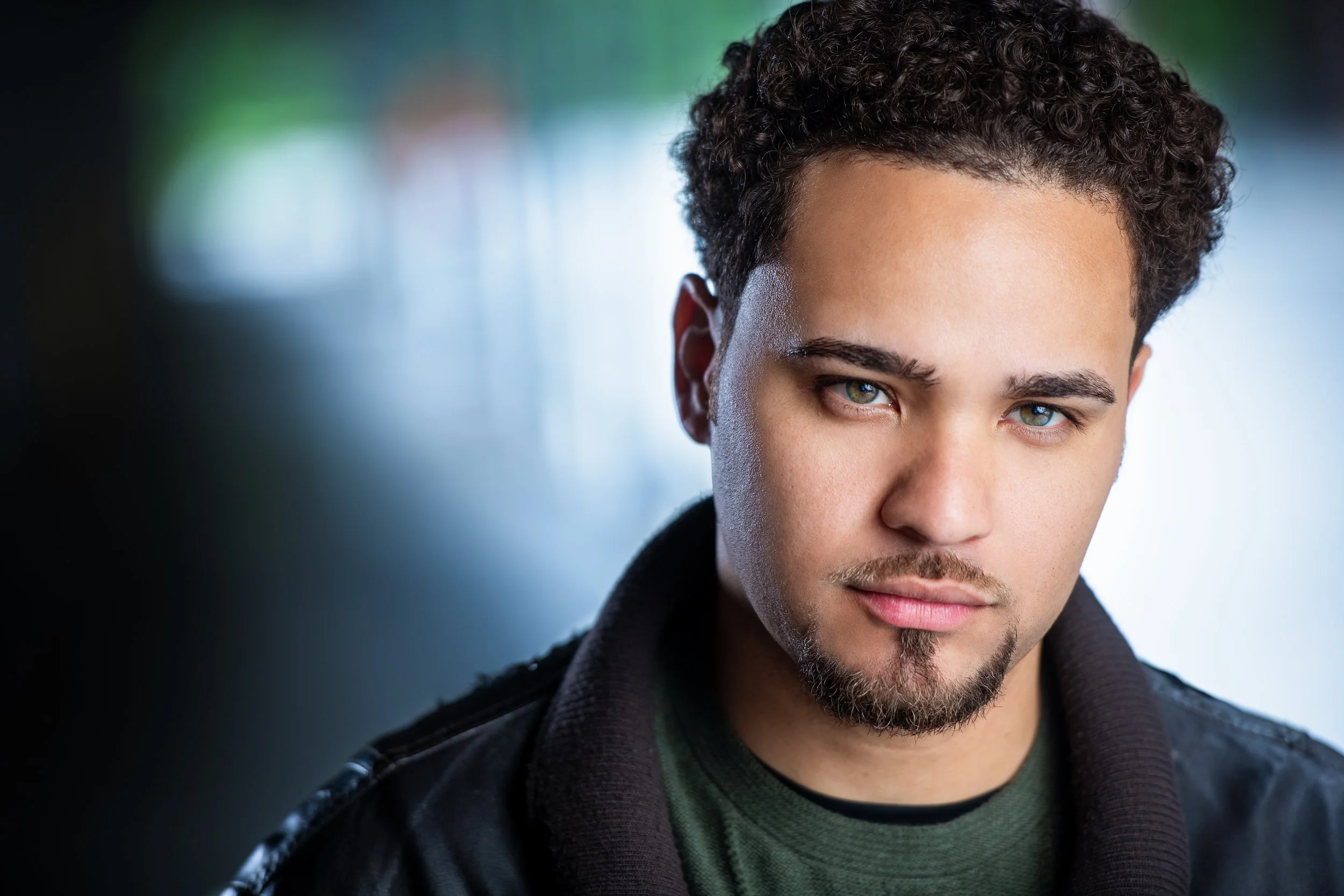 Close-up of a young man with curly hair, blue eyes, and a slight smile, wearing a dark hoodie and a green shirt, with a blurred background.