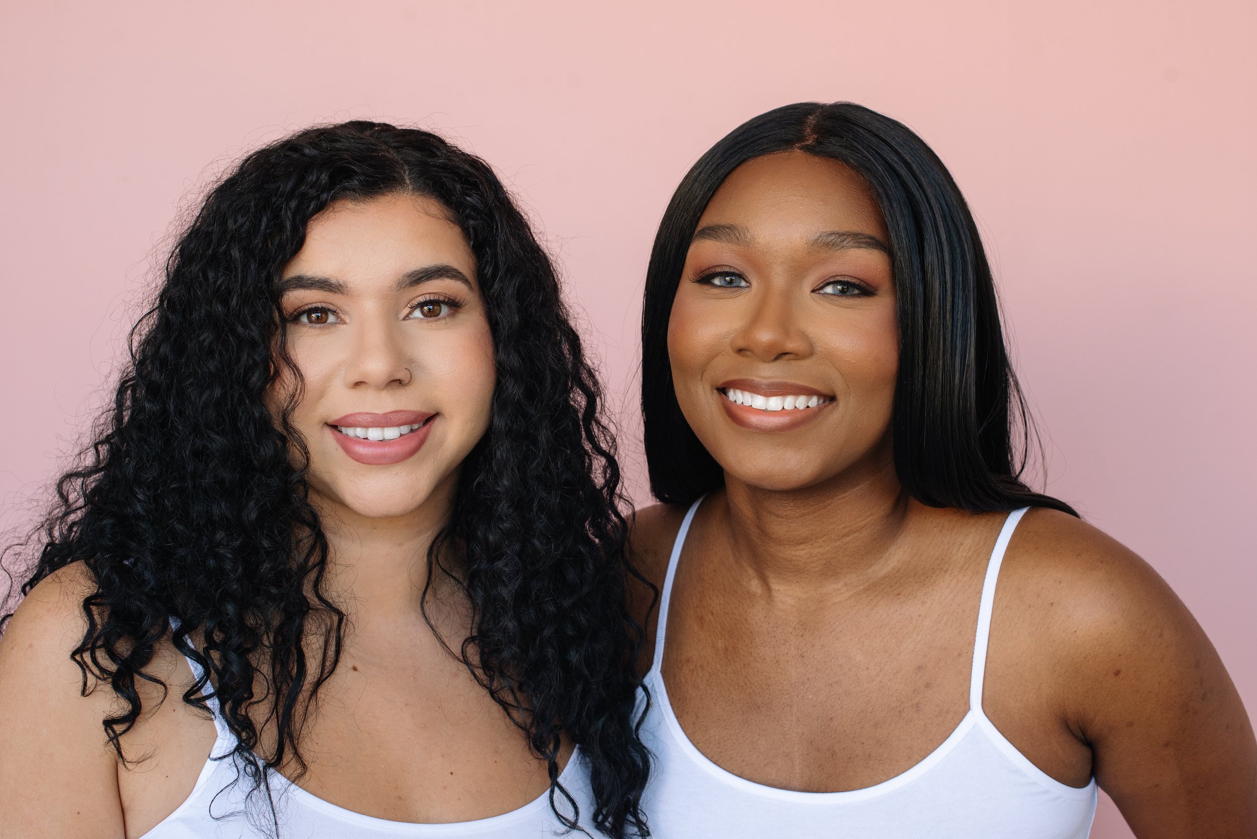 Two women standing close together, smiling, against a light pink background. Both women have dark hair, one with curly hair and the other with straight hair, wearing white tank tops.