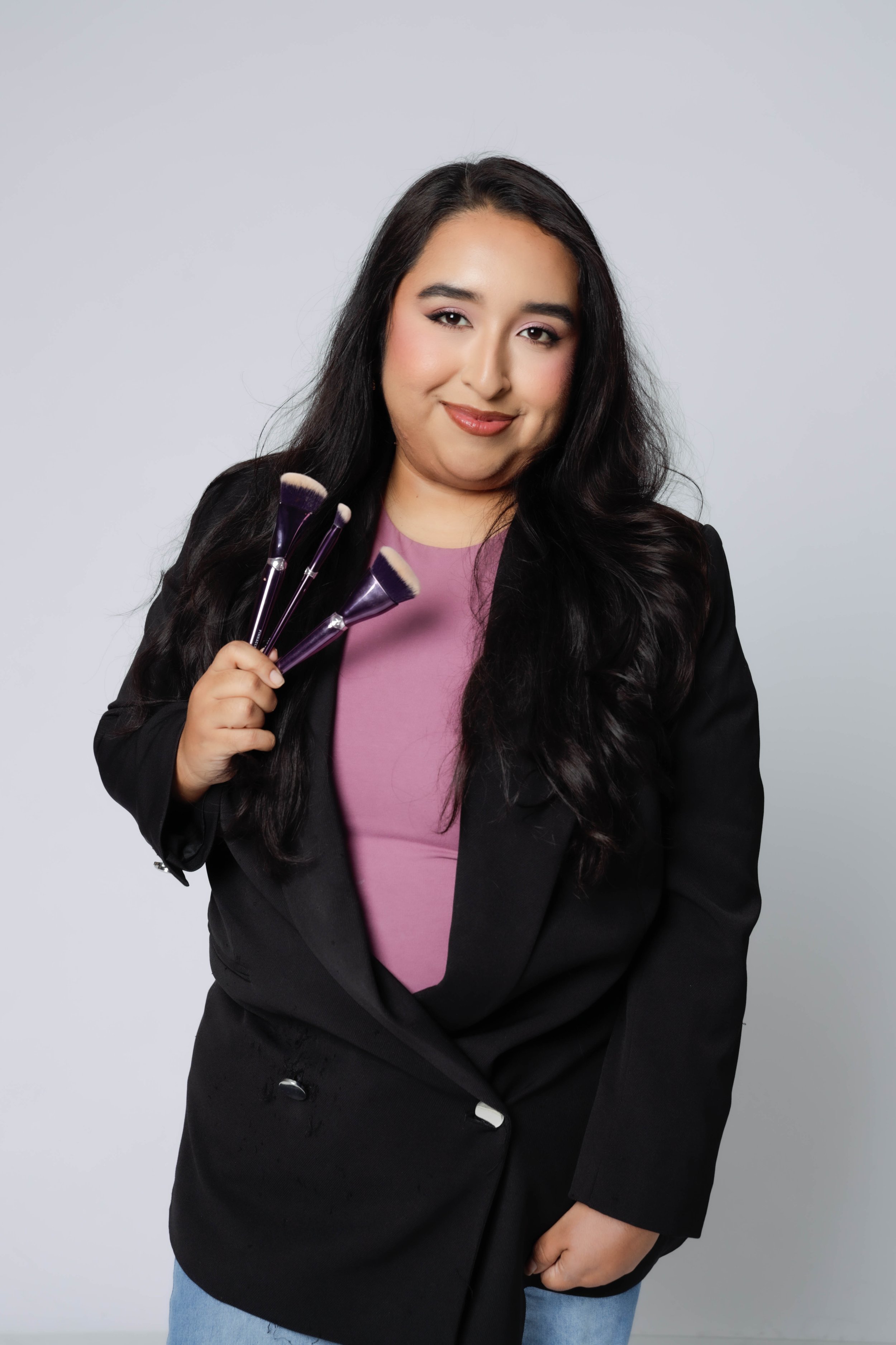 A woman with long dark hair wearing a black blazer with silver buttons, a pink top, and blue jeans, holding four makeup brushes. She is smiling and standing against a plain light grey background.