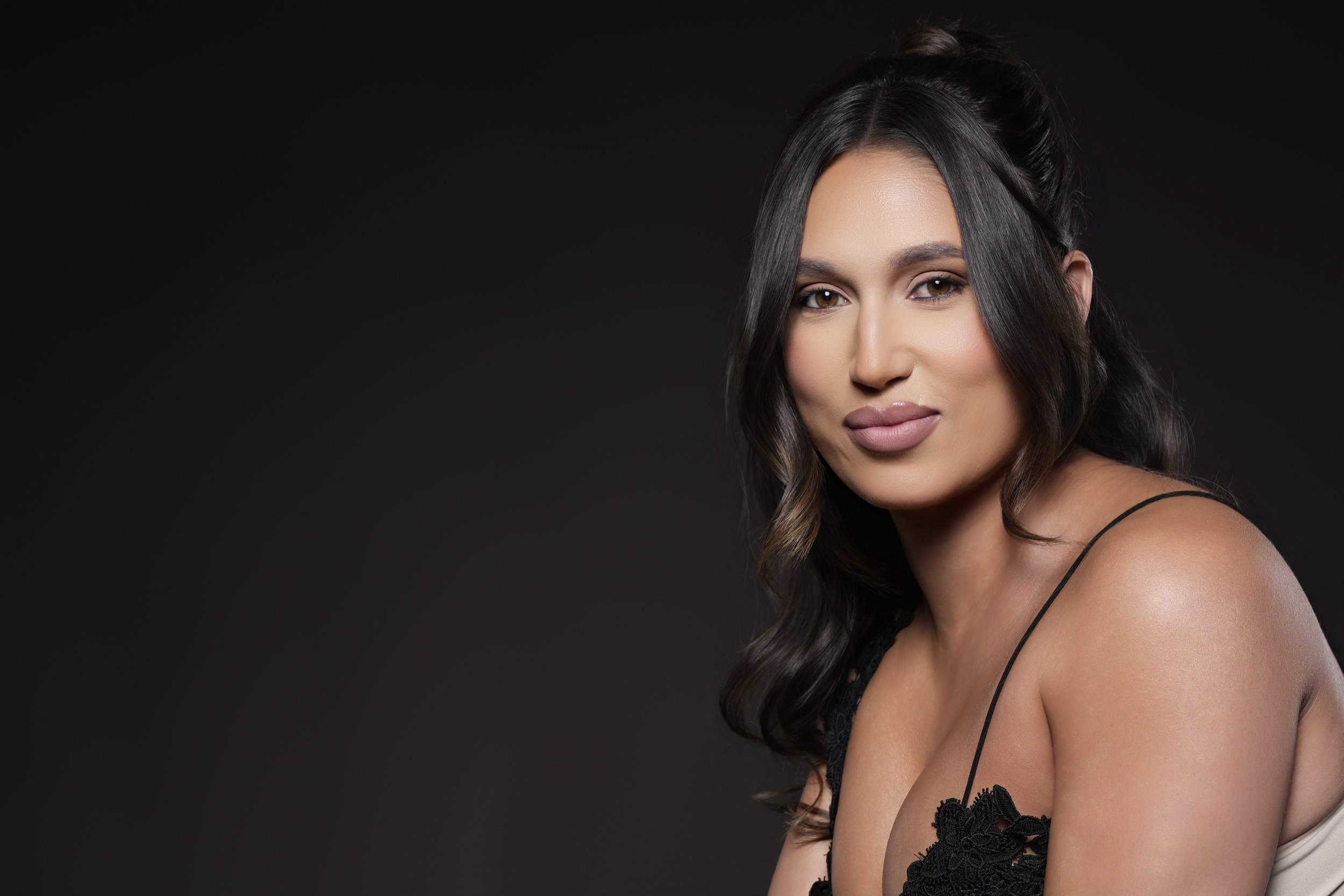 Close-up of a woman with long dark hair, wearing black lace clothing, against a black background.