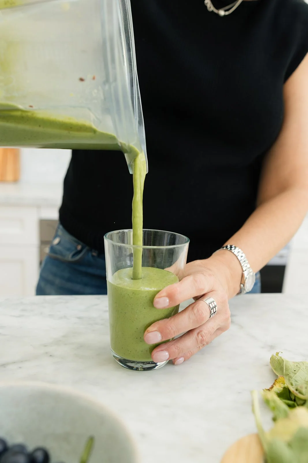 Person pouring green smoothie into a glass from a blender in a kitchen.