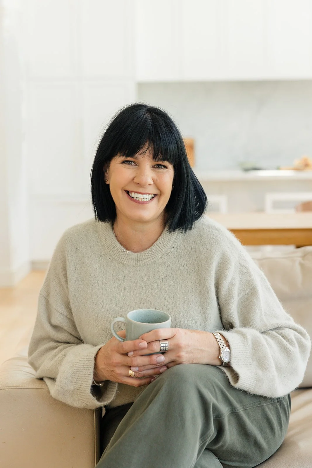 Susie the founder of Turnip Nutrition with black hair and a beige sweater sitting on a couch, smiling and holding a mug in her hands.