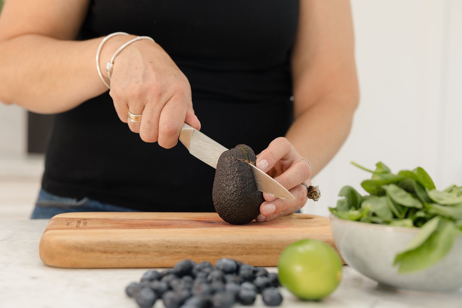 Person slicing an avocado on a wooden cutting board with a knife, with a bowl of greens, a lime, and blueberries in the foreground.