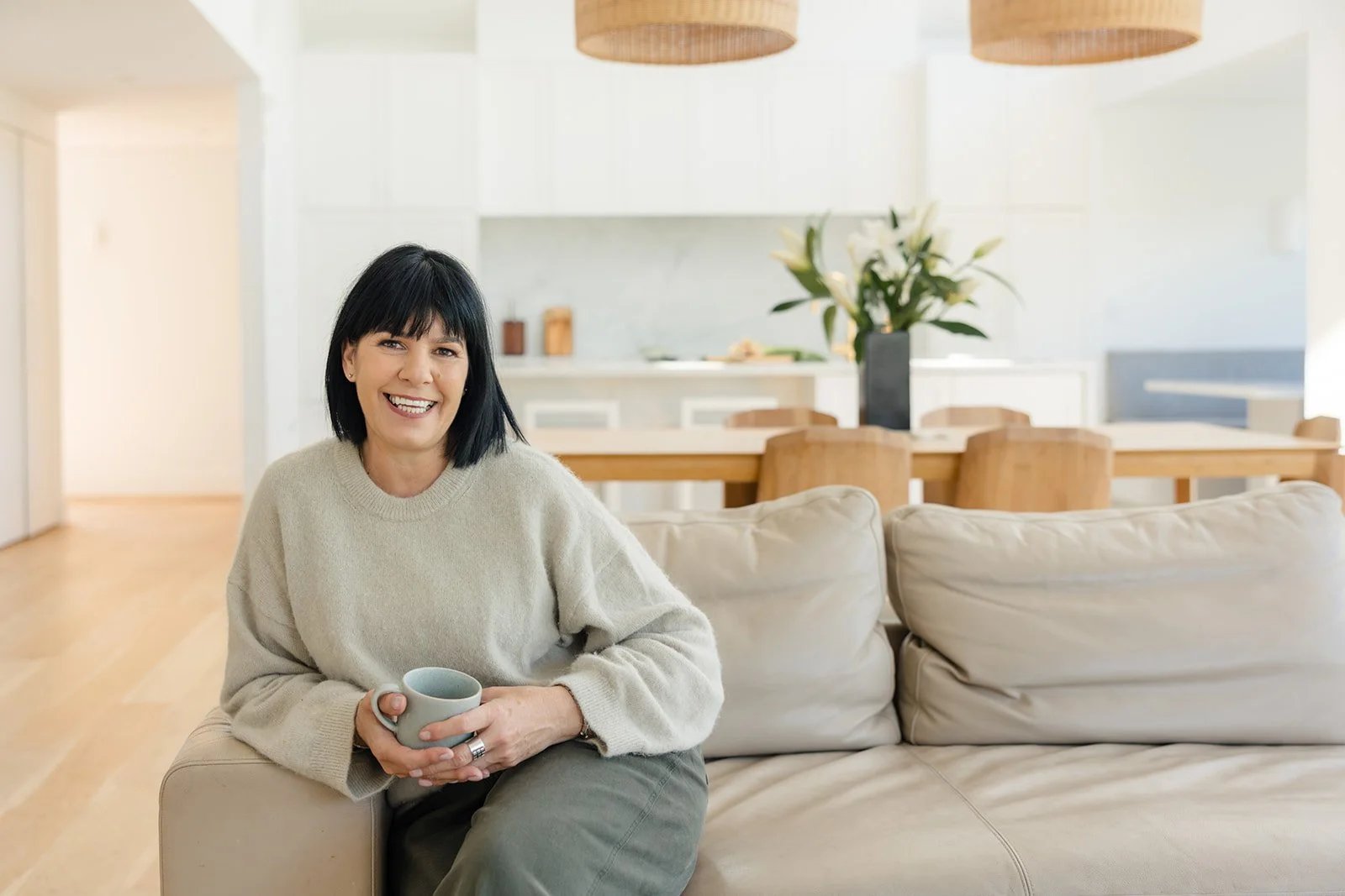 A smiling Susie, the founder of Turnip Nutrition with black hair, wearing a beige sweater, sitting on a cream-colored sofa in a bright, modern living room holding a light green mug.