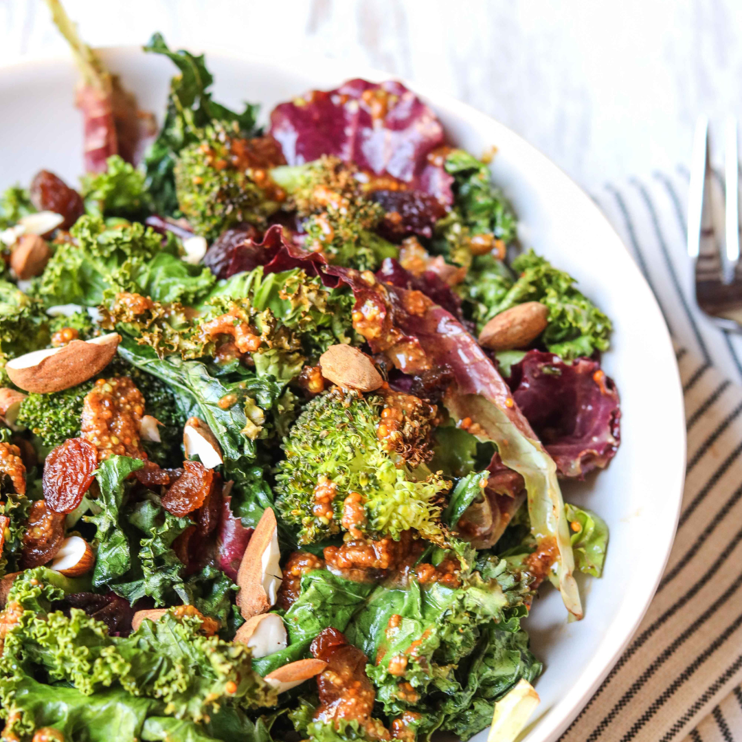 Close-up of a fresh mixed green salad with broccoli, leafy greens, toasted almonds, and a vinaigrette dressing in a white bowl on a striped napkin.