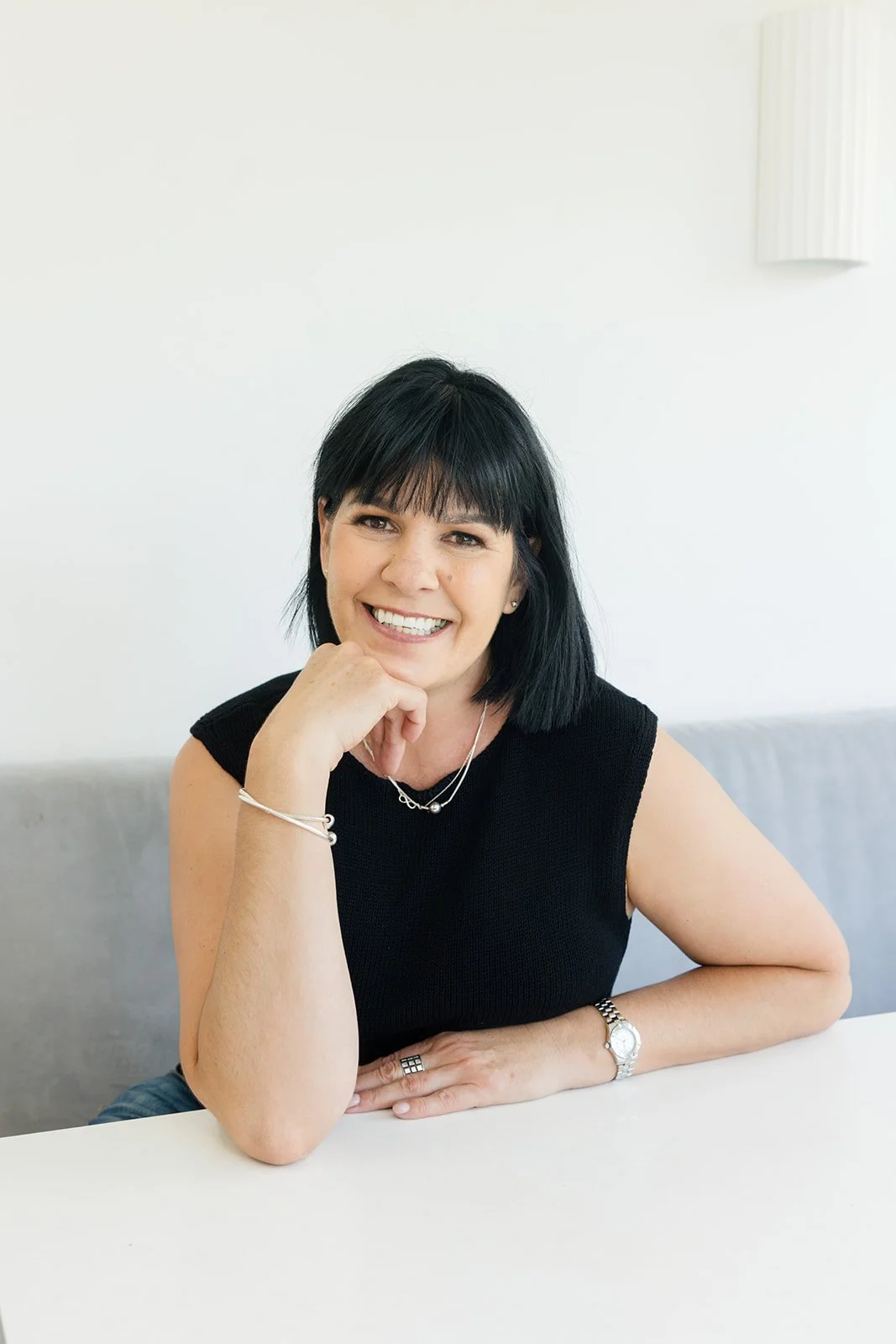 Smiling Susie, Founder of Turnip Nutrition, with black hair, wearing a black sleeveless top, jewelry, sitting at a white table in a minimalistic room with white walls.
