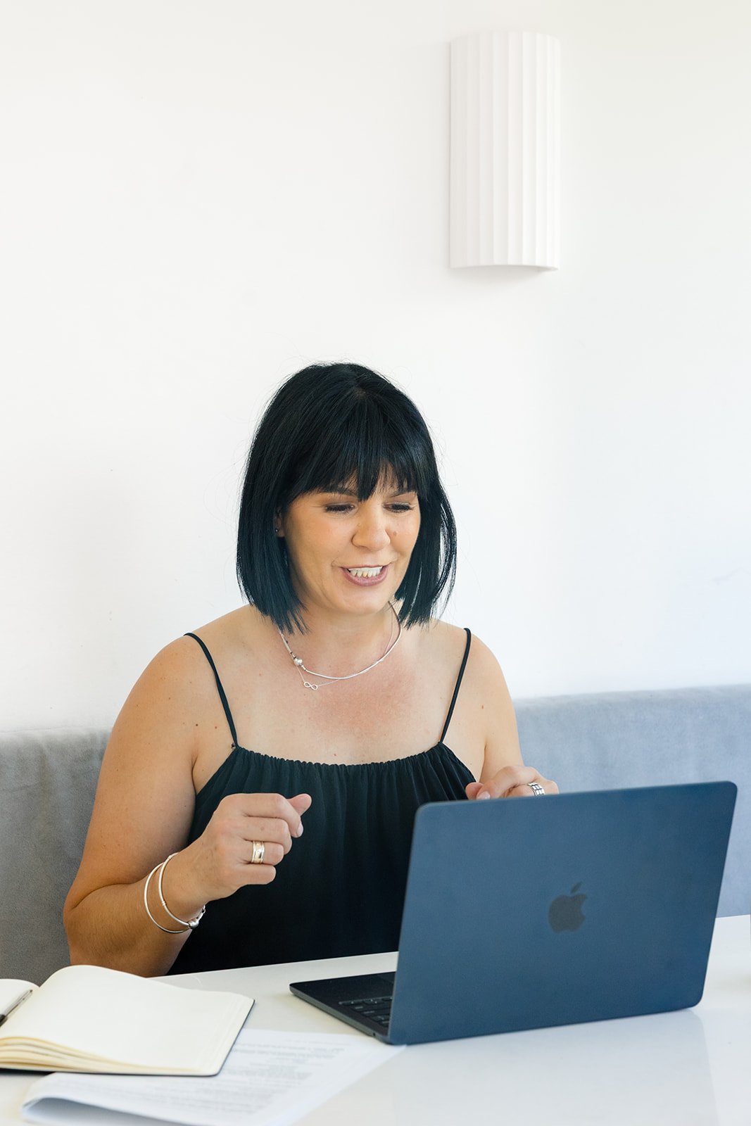 Susie, the founder of Turnip Nutrition, with black hair, wearing black spaghetti strap top, sitting at a white table with a laptop and open notebook, smiling and looking at the screen in a bright room with white walls.