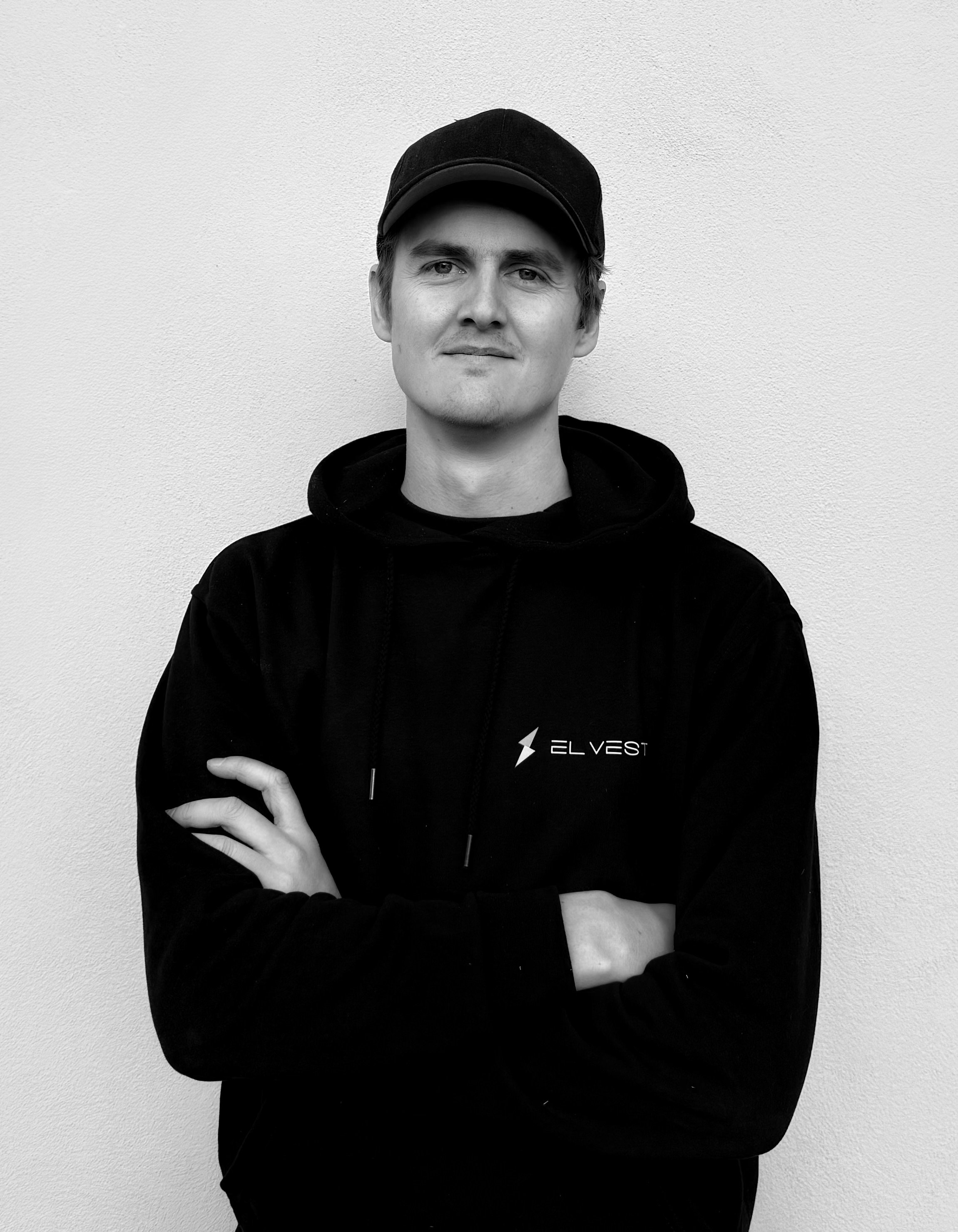 Person in black hoodie and cap standing with arms crossed against a white wall.