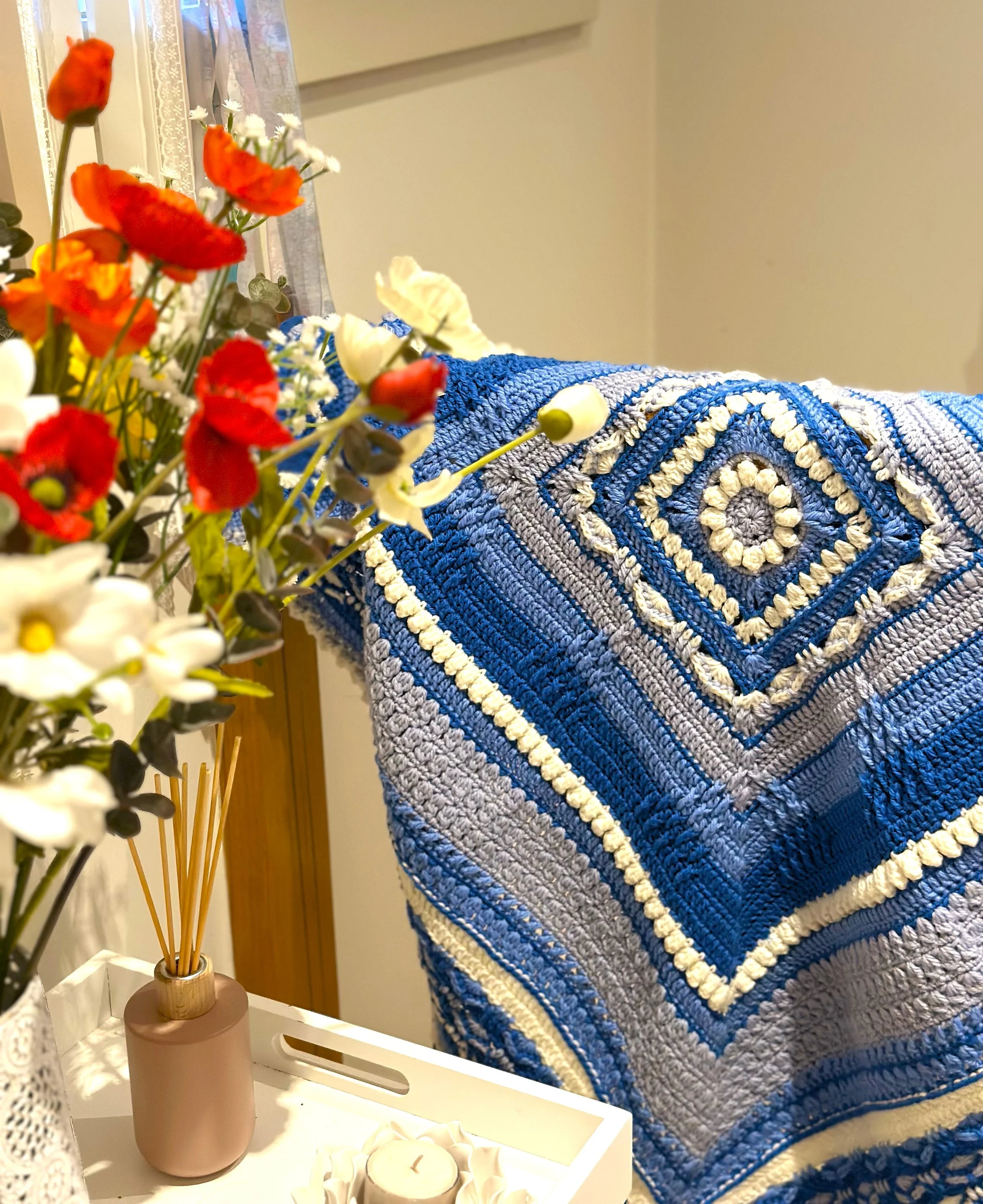 Colorful crochet blanket hanging with red and white flowers and a candle holder on a tray.