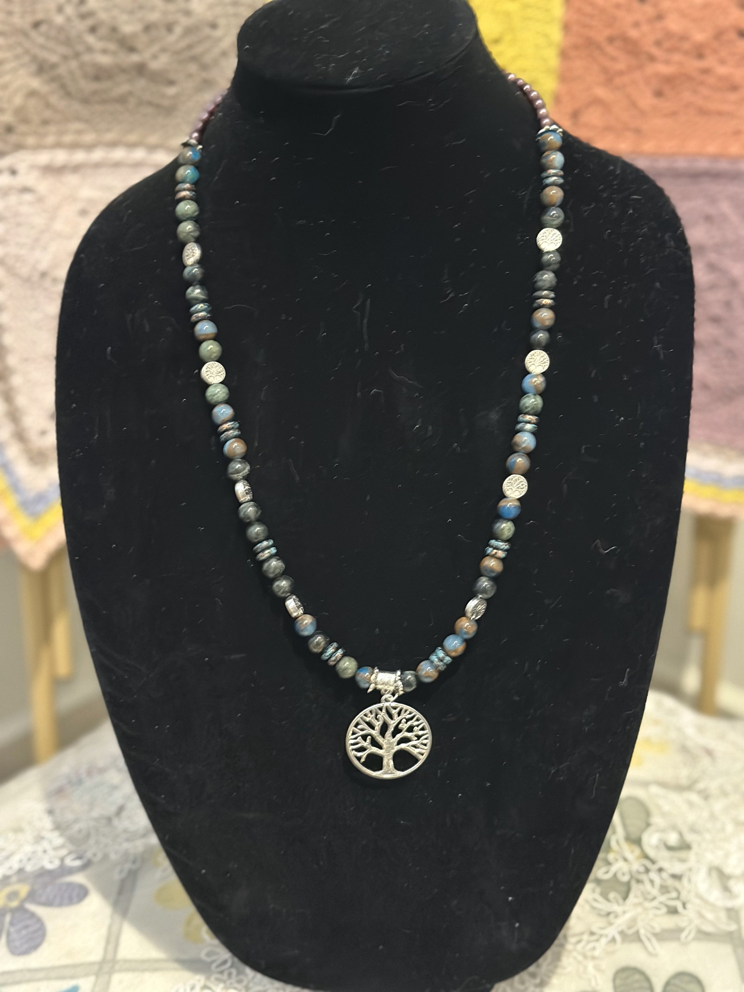 A beaded necklace with a silver tree of life pendant on a black velvet display stand.