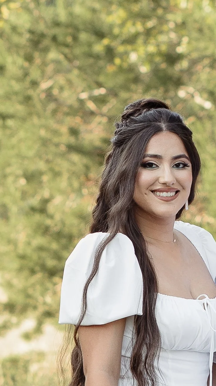 A young woman with long dark hair and a bright smile standing outdoors with blurred green trees in the background, wearing a white dress with puffed sleeves.
