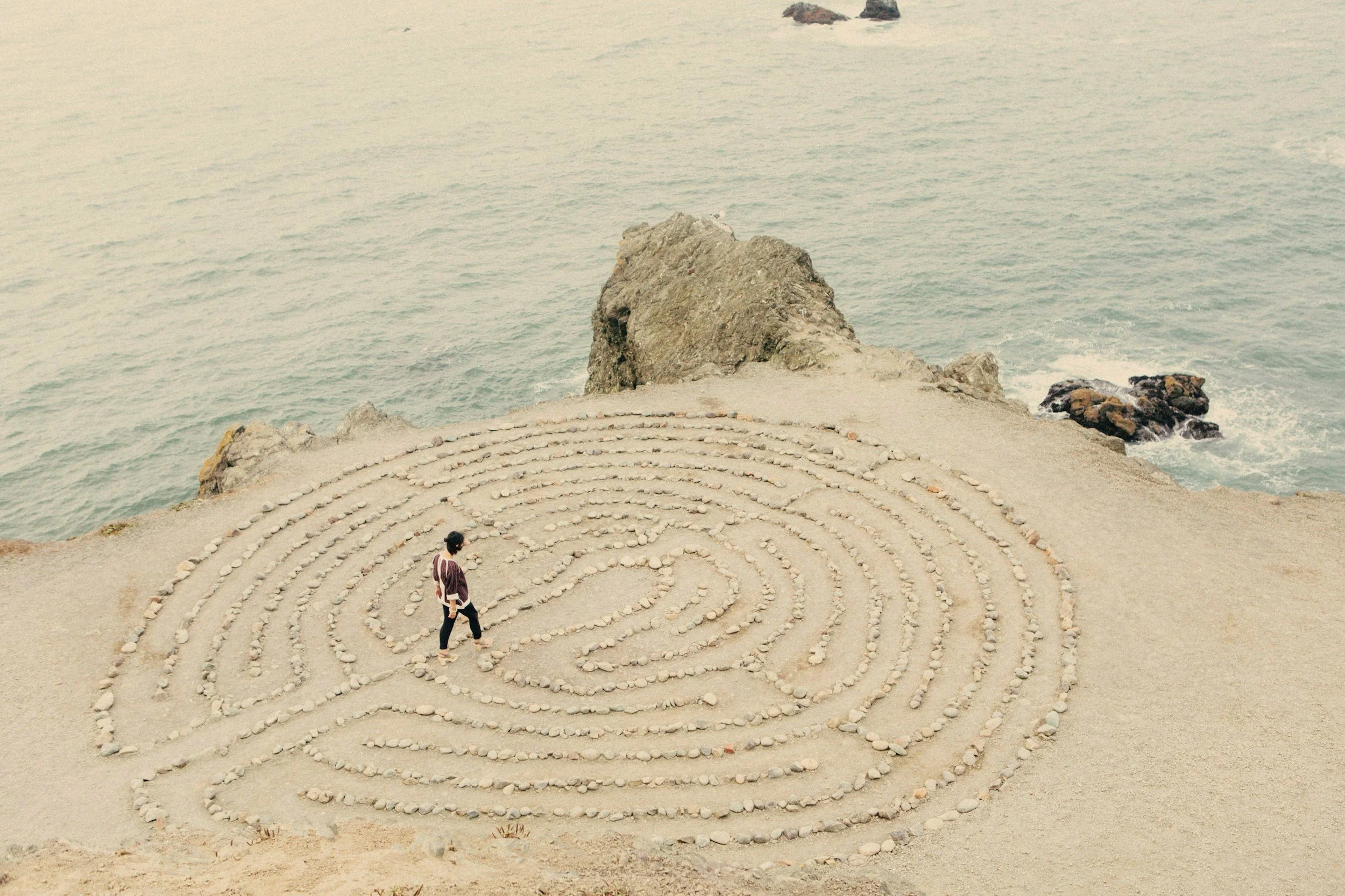 A person walking on a sandy beach with a spiral-shaped pattern made of rocks, near the ocean with large rocks in the water.