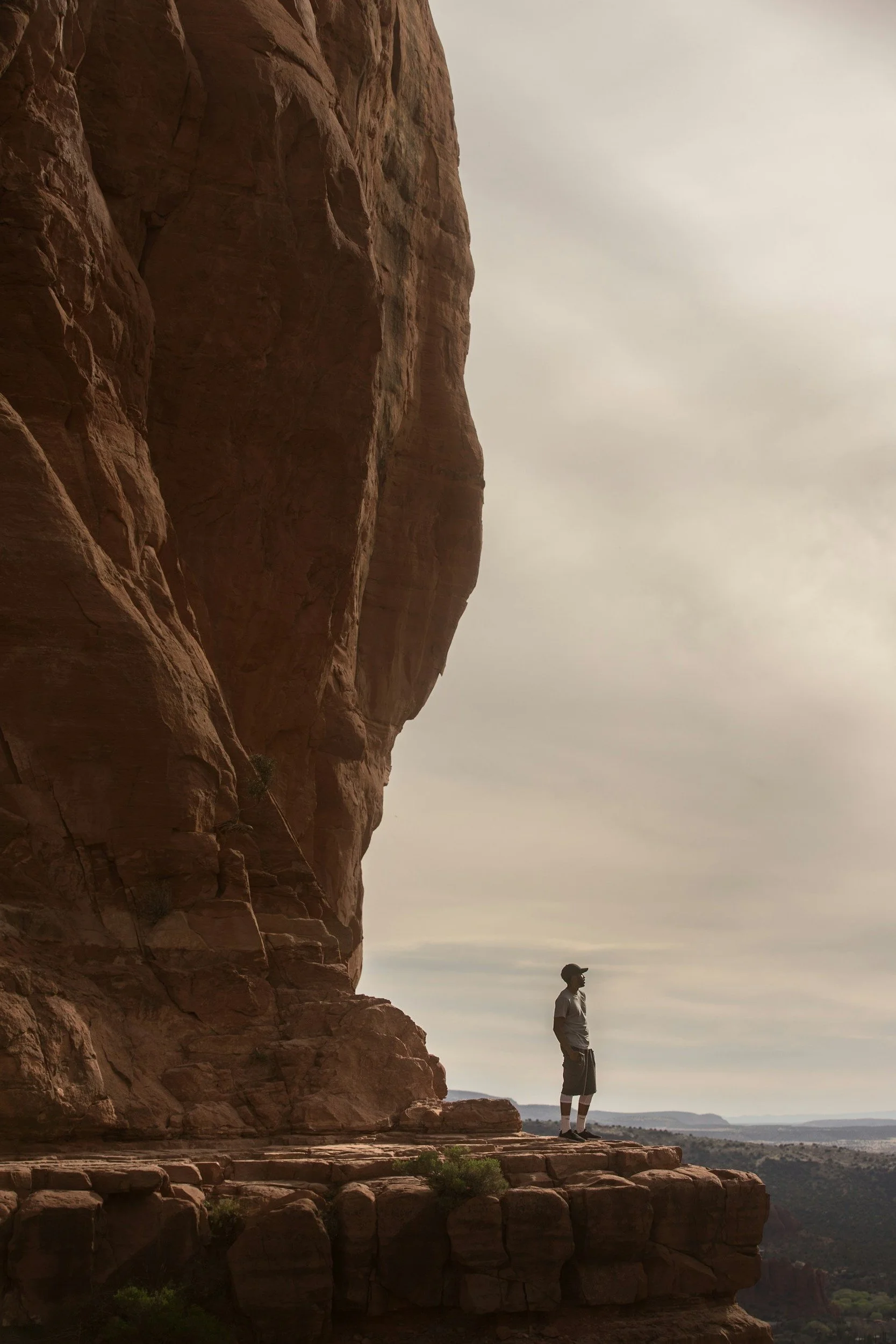 A person standing on a stone ledge next to a large, reddish-brown cliff during overcast weather, with a distant view of the landscape and a cloudy sky.