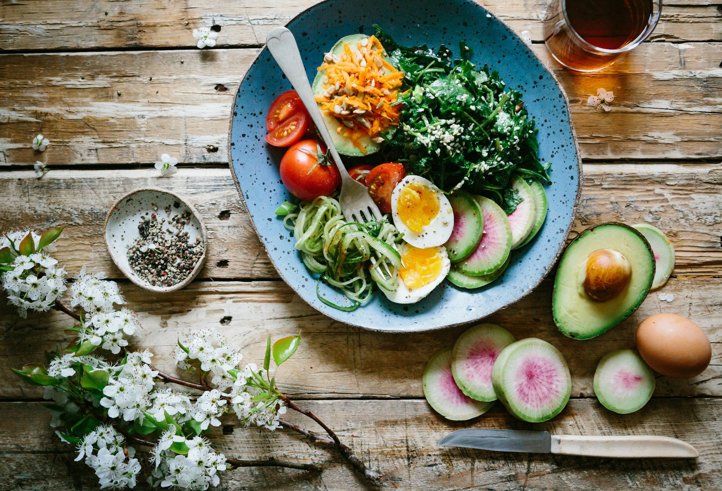 A blue plate with a salad containing tomatoes, boiled eggs, cucumber, carrots, spinach, and sesame seeds, on a rustic wooden table. A glass of tea, a knife, a small bowl of mixed peppercorns, and sliced radishes and avocado are also on the table.
