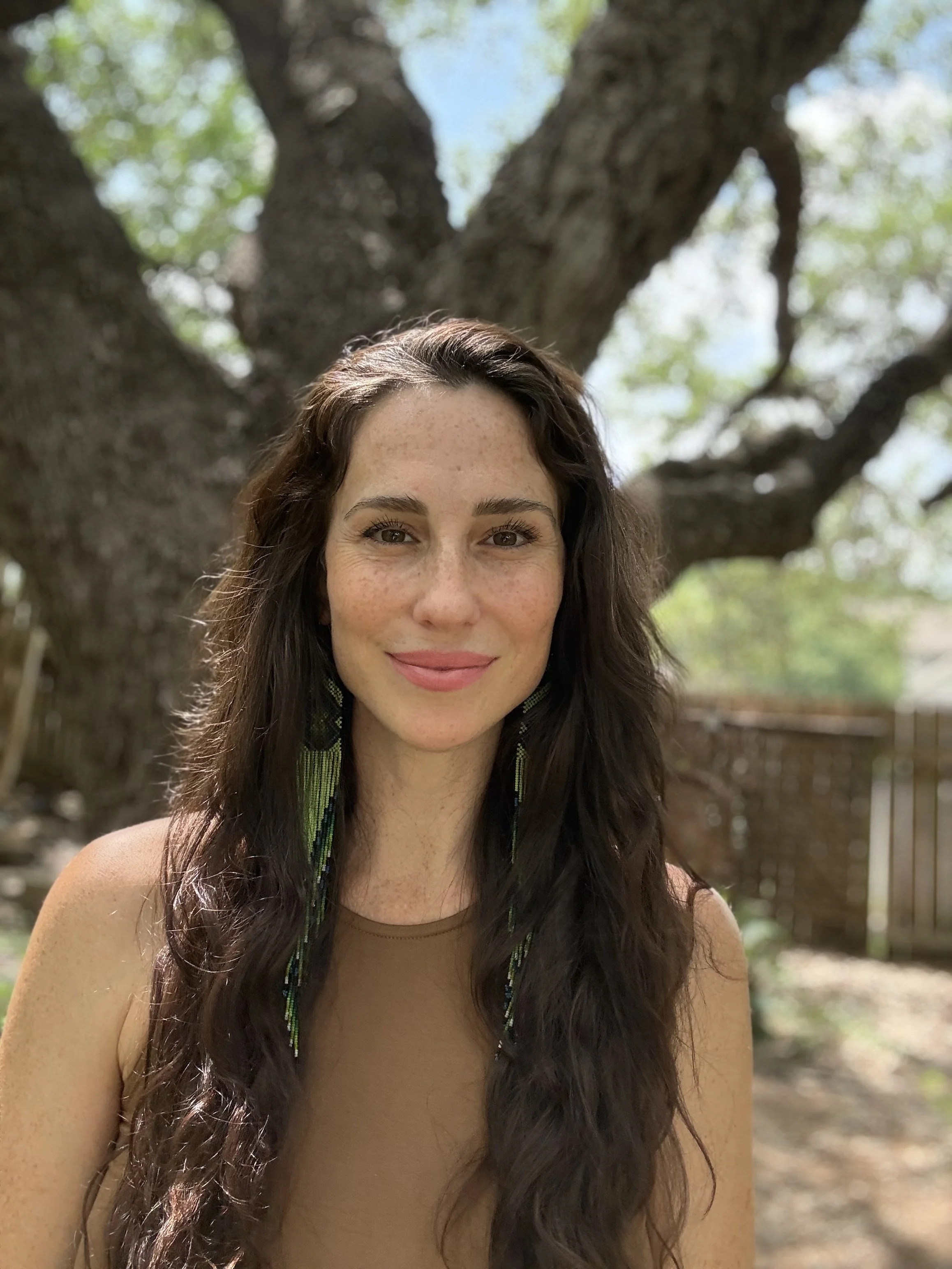 A woman with long dark hair, freckles, and a gentle smile posing outdoors with a large tree and greenery in the background.