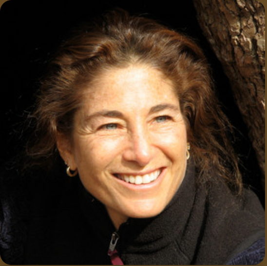 A smiling woman with light brown hair and blue eyes, wearing a black jacket and gold hoop earrings, outdoors near a tree.